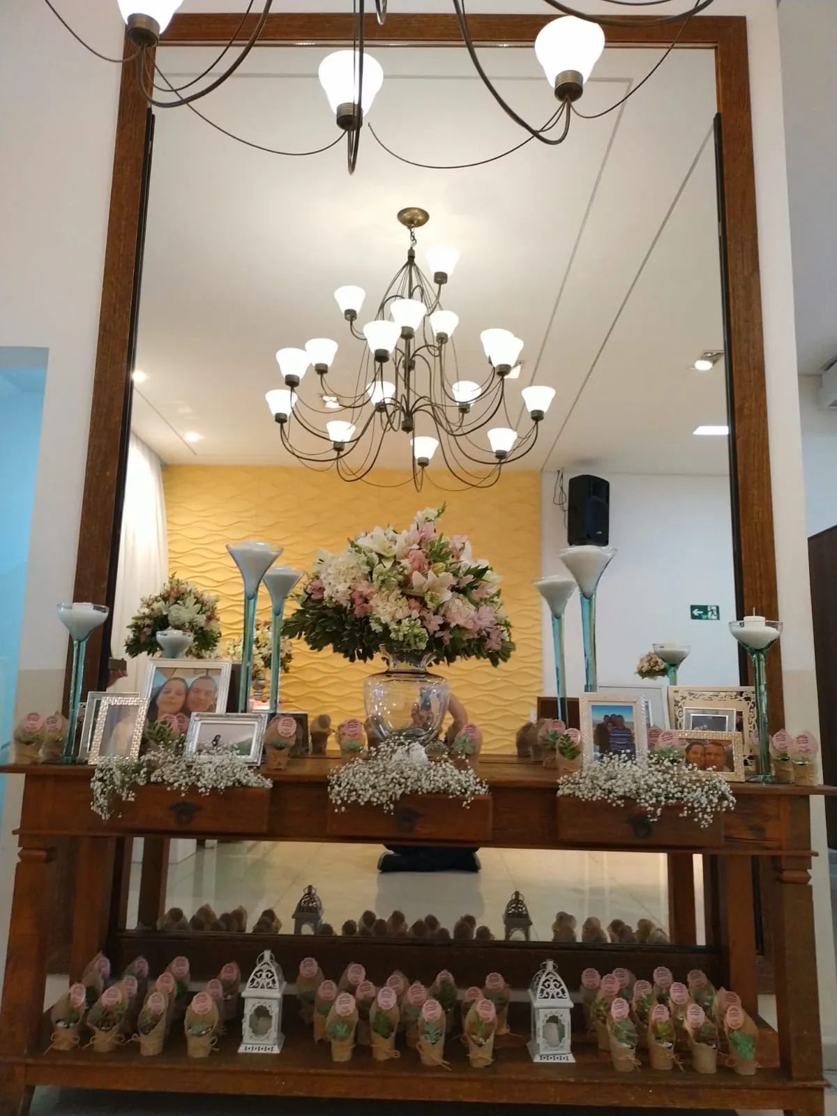Aparador de madeira com espelho, arranjo de flores e lembrancinhas na entrada do salão