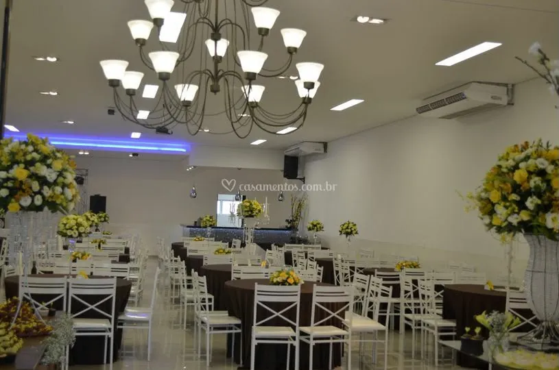 Mesas retangulares postas sob o lustre central do salão, com arranjos de flores